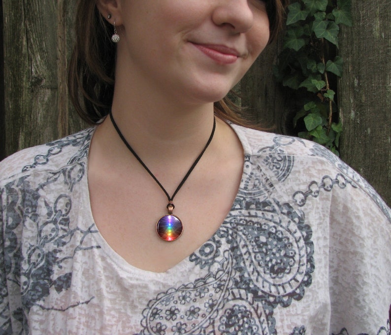 May include: A woman wearing a white shirt with a paisley pattern is wearing a necklace with a round pendant. The pendant is made of copper and has a rainbow-colored glass insert.