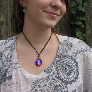 May include: A round pendant necklace featuring a vibrant, multi-colored galaxy-inspired design in shades of purple, blue, and red. The circular glass cabochon is set in a copper-toned metal frame and hangs from a simple, thin black cord.
