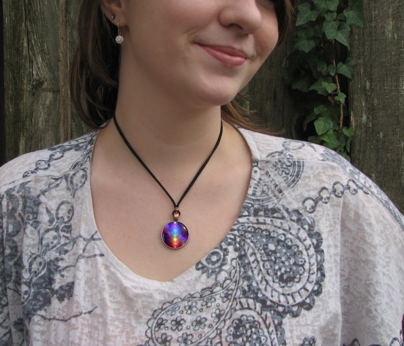 May include: A round pendant necklace featuring a vibrant, multi-colored galaxy-inspired design in shades of purple, blue, and red. The circular glass cabochon is set in a copper-toned metal frame and hangs from a simple, thin black cord.
