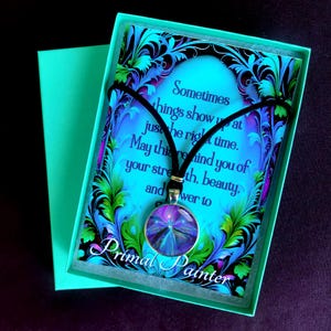 Könnte beinhalten: Eine runde Anhängerkette mit schwarzer Kordel und einem violett-blauen abstrakten Kunsteinsatz. Sie wird auf einer Karte mit dem Text "Sometimes things show up at just the right time. May this remind you of your strength, beauty, and power to grow" und "Primal Painter" in einer türkisfarbenen Geschenkbox präsentiert.