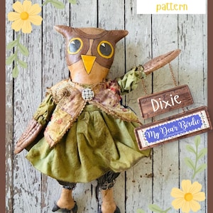 May include: A fabric owl doll with brown and tan features, wearing a green dress and a patterned vest with a button. The owl holds two hanging wooden signs that read Dixie and My Dear Birdie. The text A primitive pattern is visible.