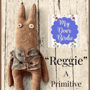 May include: A handmade brown fabric rabbit doll with button eyes and a stitched face. The doll wears a gray vest and is labeled "Reggie" with "A Primitive Rabbit Pattern" text.