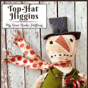 以下が含まれることがあります： シルクハットをかぶり、安全ピンで留められた緑色のベストと赤と白のドット柄のスカーフを身に着けた手作りの雪だるまの人形。人形は縫い付けられた顔とニンジンの鼻をしています。「Top-Hat Higgins」と「My Dear Birdie Patterns」の文字が見えます。
