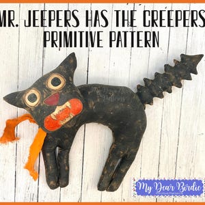 Könnte beinhalten: Eine schwarze Stoffkatzenpuppe mit abgenutztem Finish, großen cremefarbenen Augen, einer roten Nase und einem orangefarbenen Mund mit weißen Zähnen. Sie hat einen stacheligen Schwanz, ein orangefarbenes Stoffband am Hals und den Text MR. JEEPERS HAS THE CREEPERS PRIMITIVE PATTERN.