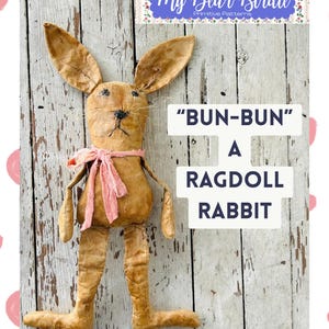 May include: A handmade ragdoll rabbit named "Bun-Bun" with long ears, a pink scarf, and stitched facial features. The rabbit is made of tan fabric and has long, floppy legs. The background is a weathered, white wooden surface.