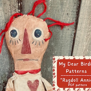 May include: Handmade ragdoll with a painted face, red yarn hair, and a red heart on the chest. The doll has large blue eyes and a red nose. The image includes a red and white sign that says "My Dear Birdie Patterns" and "Ragdoll Annie" PDF pattern.