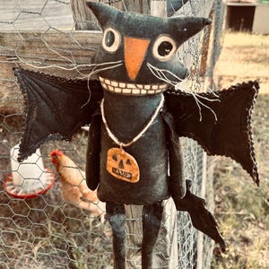 May include: A black Halloween-themed bat doll with a smiling face, orange nose, and large white eyes. The doll has black wings and a pumpkin pendant. It is hanging on a wire fence with a chicken coop in the background.