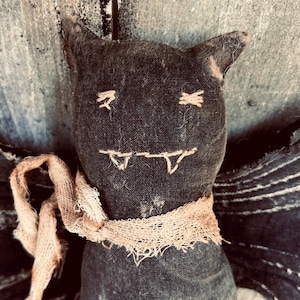May include: A handmade, dark gray bat doll with stitched facial features and a tan burlap scarf. The bat has pointed ears and stitched eyes and fangs. The wings are visible on the sides. The doll is made of a textured fabric.