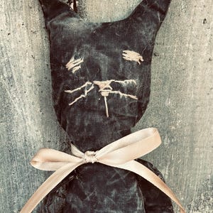 May include: A handcrafted black cat doll with stitched facial details and a cream ribbon bow. The cat features pointed ears and a distressed, vintage look. The doll is made of fabric, giving it a rustic, handcrafted appearance.