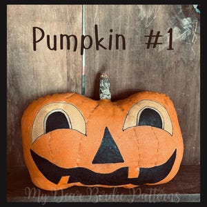 May include: A handmade orange fabric pumpkin with a carved jack-o'-lantern face. The pumpkin features black eyes, a triangular nose, and a smiling mouth. The text "Pumpkin #1" is above the pumpkin. The words "My Dear Birdie Patterns" are at the bottom.