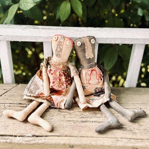 May include: Two handcrafted rag dolls with button eyes and stitched details. One doll is dressed in a floral print dress, the other in a beige apron. Both dolls have long limbs and sit together on a wooden surface, giving a vintage, folk art feel.