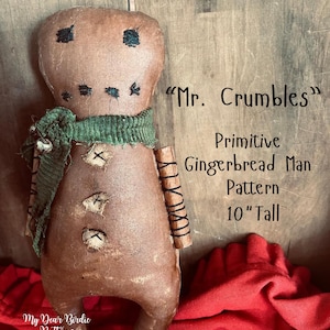 May include: A handmade gingerbread man doll, approximately 25 cm tall, with a distressed brown finish. It features black stitched eyes and mouth, a green scarf, and stitched details. The text "Mr. Crumbles" and "Primitive Gingerbread Man Pattern" are visible.