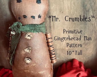 Può includere: Bambola fatta a mano a forma di omino di pan di zenzero, alta circa 25 cm, con finitura marrone anticata. Presenta occhi e bocca cuciti in nero, una sciarpa verde e dettagli cuciti. Il testo "Mr. Crumbles" e "Primitive Gingerbread Man Pattern" sono visibili.