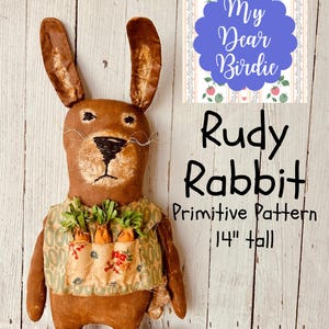 May include: A brown fabric rabbit doll named "Rudy Rabbit" with long ears and a vest holding carrot decorations. The doll is 14 inches tall. The image includes the text "My Dear Birdie" and "Primitive Pattern".