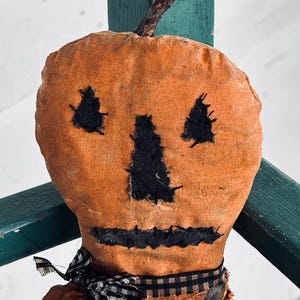 May include: A handmade orange fabric pumpkin doll with a stitched black face and a twig stem. The doll has a black and white checkered ribbon around its neck, evoking a rustic, vintage Halloween theme.