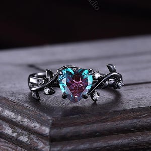 Könnte beinhalten: Ein schwarzer Ring mit einem herzförmigen, farbwechselnden Edelstein. Der Ring hat ein weinrebenartiges Design mit kleinen weißen Steinen und liegt auf einer dunklen Holzoberfläche. Der Edelstein wechselt von Türkis zu Lila.