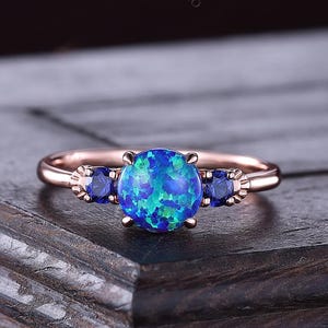 May include: A rose gold ring featuring a large, round, blue opal center stone with vibrant green and blue hues. Two smaller blue stones flank the center stone. The ring is set on a dark wooden surface. The text "ConnieJewelsStudio" is visible.