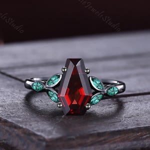May include: A black metal ring featuring a central coffin-shaped red gemstone. The band is adorned with small, marquise-cut green stones arranged in a leaf-like pattern on both sides of the central setting, creating a symmetrical design.