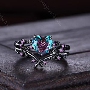 May include: A black metal ring featuring a central heart-shaped color-changing stone that shifts between blue and purple. The band is designed with a vine-like motif accented with small, round, pink stones embedded along the metal branches.