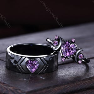May include: Two matching rings. One is a black band with a heart-shaped purple gem. The other ring has a heart-shaped purple gem with crescent moon accents and smaller purple gems. The rings are on a dark wooden surface.