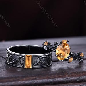 Op de afbeelding: Twee ringen met gele edelstenen. Eén ring heeft een rechthoekige steen in een band met bladdetails. De andere ring heeft een ovale steen op een zwart takontwerp met kleine gele accenten. De ringen worden getoond op een donker houten oppervlak.