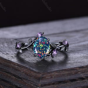 May include: A black ring with an oval, iridescent opal centre stone. The ring features leaf and amethyst accents. The ring is set on a dark wooden surface. The text "ConnieJewelsStudio" is visible in the background.