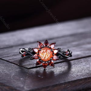 May include: A black metal ring featuring a central round orange sunstone surrounded by a halo of marquise-cut red garnet stones. The band has a twisted vine-like design with small black metal leaves extending from the sides of the central setting.