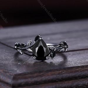 Puede incluir: Anillo negro con una piedra preciosa en forma de pera y diseño floral. El anillo tiene una banda de metal oscuro con detalles de hojas y vides. El anillo está sobre una superficie de madera oscura. La piedra es negra.