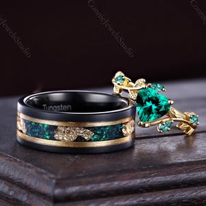 May include: Two rings: a black tungsten band with gold and green inlay, and a gold ring with a heart-shaped emerald and small emerald accents. The rings are displayed on a dark wooden surface, showcasing the intricate details of the jewelry.