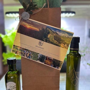 May include: A gift set featuring two green glass bottles of olive oil, a brown paper bag with green tissue paper, and a card with the word "Fratelli" and a landscape photo. The bottles have olive branch designs and labels.