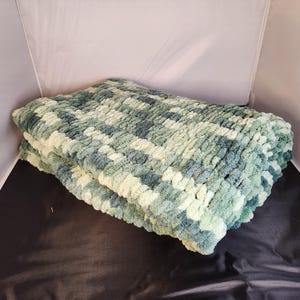 May include: A folded, chunky knit blanket in shades of green and cream. The blanket has a textured, plush appearance, with a pattern of alternating colors. The soft blanket is ideal for home decor and comfort.