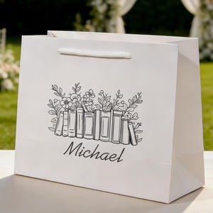 May include: White gift bag featuring a black ink illustration of books and flowers. The design includes a row of books with floral elements above. The name "Michael" is written below the illustration.