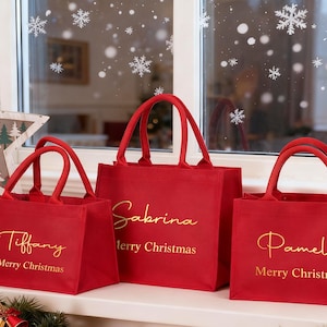 Puede incluir: Tres bolsas rojas con letras doradas. Cada bolsa tiene un nombre y "Feliz Navidad" impreso. Las bolsas están en un alféizar con decoraciones navideñas, incluyendo campanas y una estrella.