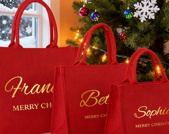 Könnte beinhalten: Drei rote Tragetaschen mit goldenen Buchstaben. Die größte Tasche trägt die Aufschrift "Fran" und "MERRY CH", die mittlere Tasche trägt die Aufschrift "Bet" und "MERRY CH", und die kleinste Tasche trägt die Aufschrift "Sophia" und "MERRY CHRISTMAS".