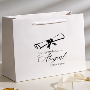 Peut inclure: Sac cadeau blanc avec une illustration de parchemin noir et le texte "Félicitations Abigial CLASS OF 2026". Le sac a une poignée en corde blanche.