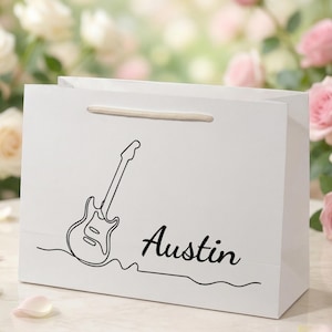 May include: White gift bag with a black line drawing of an electric guitar and the name "Austin" in script. The bag has a beige handle and is set against a blurred background of roses.