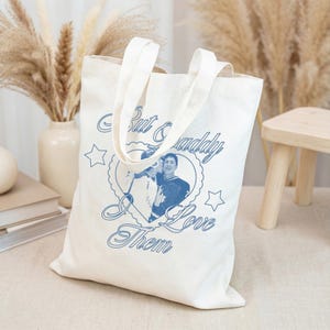 Puede incluir: Bolso tote de lona color crema con texto azul y foto. El texto dice "But & Daddy I Love Them" alrededor de una imagen en forma de corazón de dos personas. El bolso tiene dos asas.