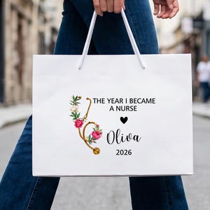 Può includere: Una borsa regalo in carta bianca con manici in corda bianca, caratterizzata da un grafico a stetoscopio dorato decorato con fiori rosa e bianchi. Il testo recita THE YEAR I BECAME A NURSE, un cuore nero, il nome Oliva e l'anno 2026.