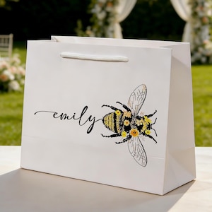 May include: White gift bag with a bee illustration and the name "emily" in cursive. The bee has yellow and black stripes and is decorated with yellow flowers. The bag has a white rope handle.