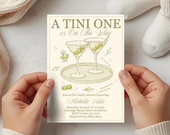 May include: Baby shower invitation with the text "A Tini One is On The Way." The invitation features two martini glasses with olives, a plate, and the name "Michelle Arlo." The invitation is held by a person.