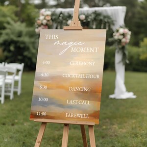 May include: A wooden easel displays a wedding schedule sign. The sign features the text "THIS magic MOMENT" and a timeline with times and event names like "CEREMONY" and "FAREWELL". The background has a sunset-like color palette.