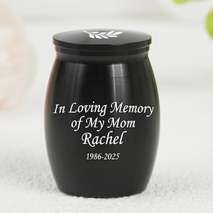 May include: Black memorial urn with a lid, inscribed with "In Loving Memory of My Mom Rachel 1986-2025." The urn has a simple design with a white leaf detail on the lid. A poignant keepsake for remembrance.