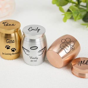 Könnte beinhalten: Vier kleine Urnen in Gold, Silber und Roségold. Die goldene Urne trägt den Namen "Gina" und Pfotenabdrücke. Die silberne Urne trägt den Namen "Emily" und Engelsflügel. Die roségoldene Urne hat einen Pfotenabdruck und den Namen "Hilda".