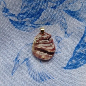 May include: A small, handcrafted pendant with a gold-colored bail. The pendant is a unique shape, with a swirling pattern of white, pink, and dark purple. The background is a light blue fabric with a floral design.