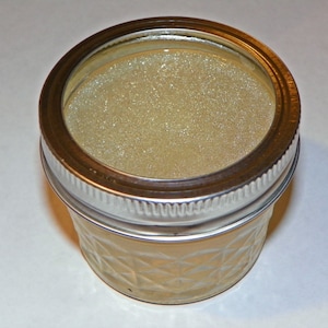 May include: A clear glass jar with a silver lid filled with a light yellow, slightly translucent substance. The substance has a grainy texture.