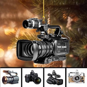 May include: A black video camera ornament with the text "YOUR NAME" on the side. The ornament hangs from a gold string. The image includes four smaller camera images at the bottom.