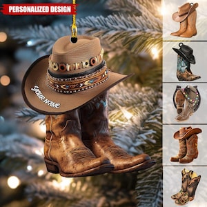 Puede incluir: Adorno navideño de sombrero y botas de vaquero marrones con el texto "your name" en el sombrero. El sombrero tiene bandas decorativas con cuentas y diseños. El adorno cuelga de un hilo dorado. Las palabras "PERSONALIZED DESIGN" están arriba.