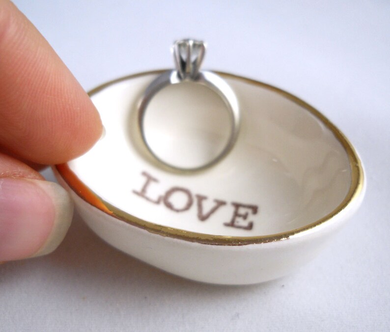 May include: White ceramic ring dish with a gold rim and the word "LOVE" printed in brown. A silver ring with a single diamond is sitting in the dish.