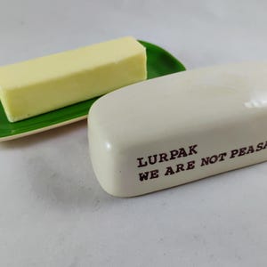 May include: A butter dish with a stick of butter on a green dish. The butter dish is off-white with the text "LURPAK WE ARE NOT PEASANTS" in brown. The butter is a light yellow color.