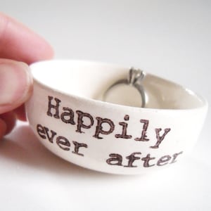 May include: White ceramic ring dish with the words "Happily ever after" printed in brown. A silver ring with a single diamond is sitting in the dish.